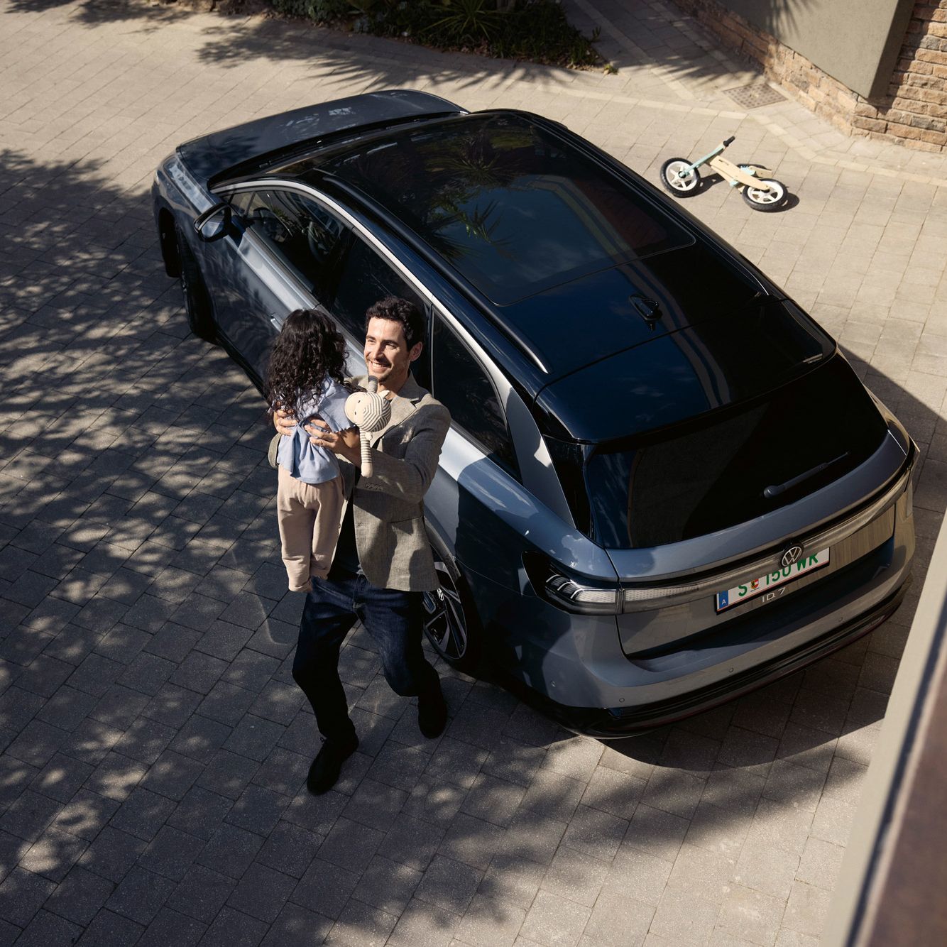 Mann hebt sein Kind hoch, was eine Puppe in der Hand halt und dahinter steht geparkt in der Einfahrt ein ID.7 Tourer und ein umgekipptes Kinderfahrrad.