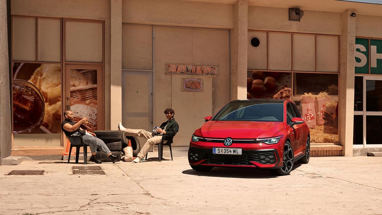 Zwei junge Männer sitzen vor einem Geschäft neben einem roten VW Golf GTI.