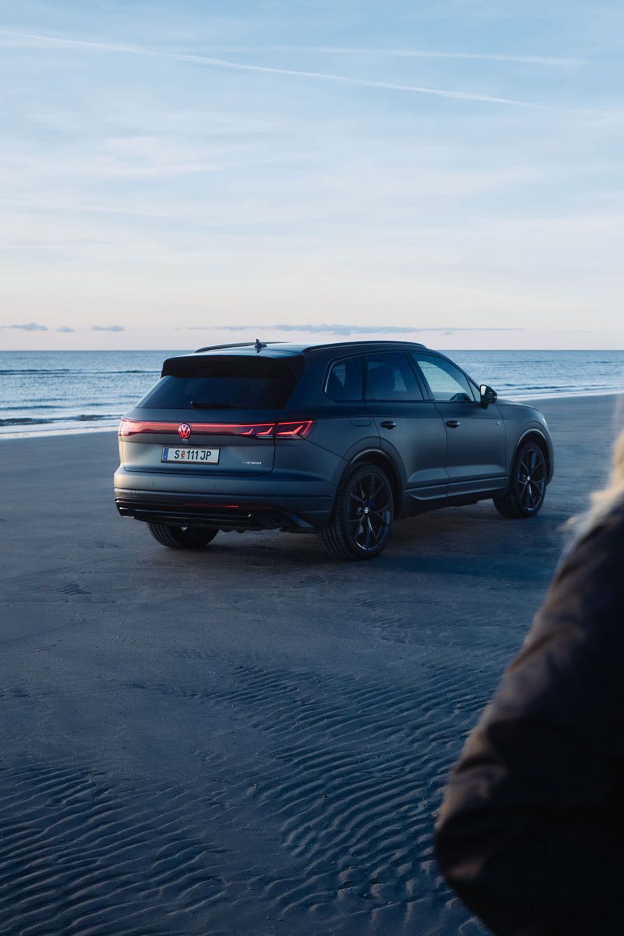 Eine Frau mit blondem Haar steht im Vordergrund an einem Strand, während ein dunkler Touareg im Hintergrund nahe dem Wasser parkt.