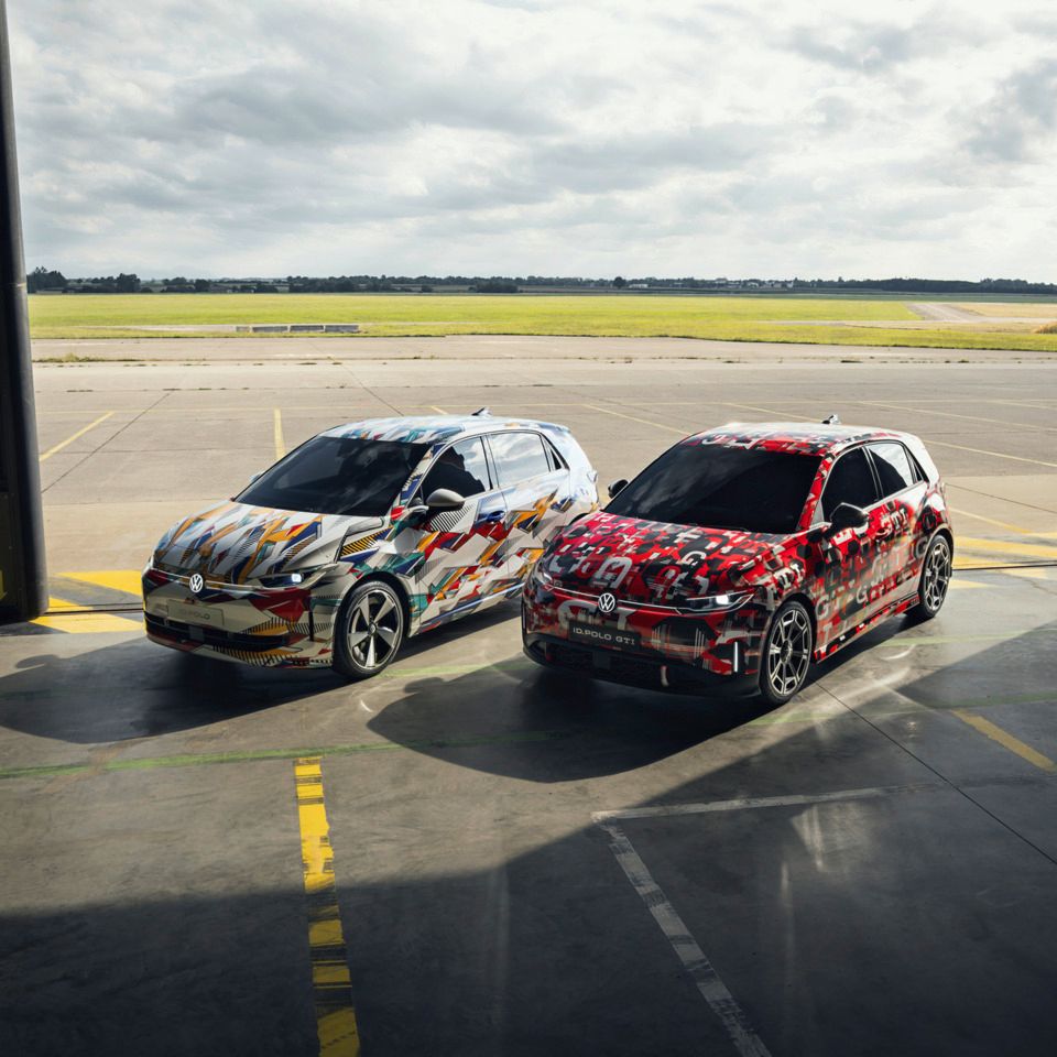 Frontale Sicht auf den ID. Polo und ID. Polo GTI. Beide Fahrzeuge sind bunt foliert und stehen in einem Hangar.