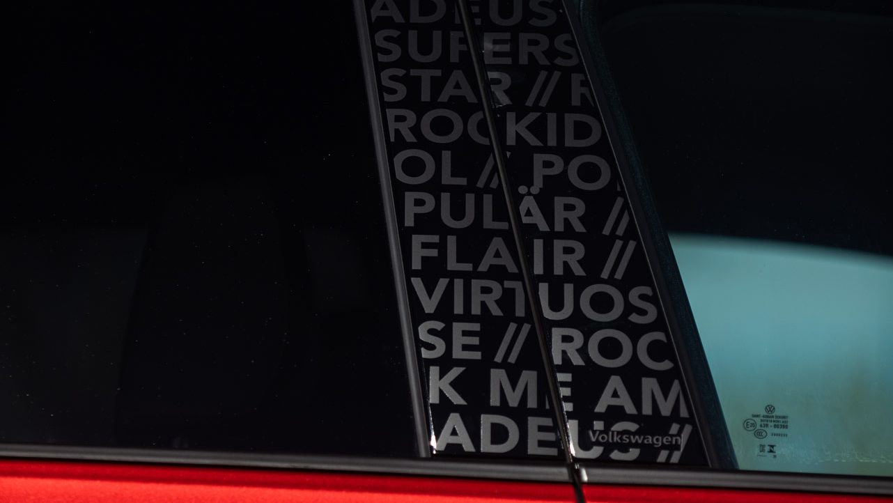 Ausschnitt eines roten Autos, aufgenommen seitlich bei Tageslicht. Durch das hintere Seitenfenster ist eine schwarze Verkleidung mit weißer, teils verdeckter Schrift in Blockbuchstaben zu sehen, unter anderem Wörtern wie „ROCK“, „POPULAR“ und „VIRTUOSO“. Im Hintergrund erkennt man unscharf blauen Himmel und etwas Baumlaub.