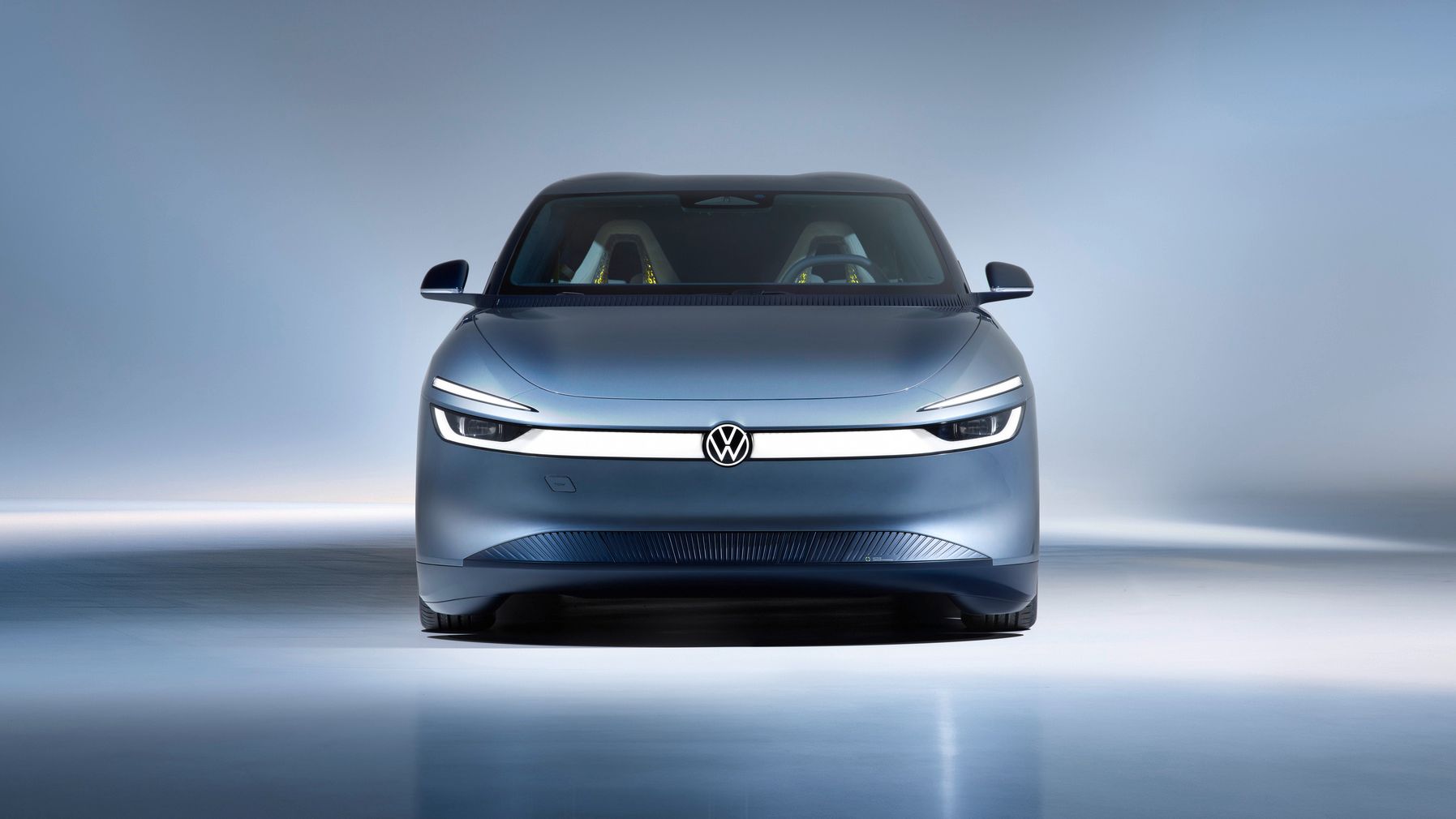 Frontansicht eines modernen, silbergrauen Elektroautos von Volkswagen in einem hell ausgeleuchteten Studio. Das Fahrzeug hat schmale, durchgehende LED-Scheinwerfer und ein minimalistisches, glattes Design. Im Innenraum sind zwei farbige Sitze schemenhaft erkennbar.