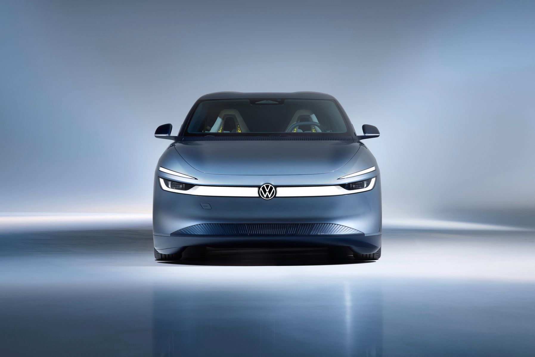 Frontansicht eines modernen, silbergrauen Elektroautos von Volkswagen in einem hell ausgeleuchteten Studio. Das Fahrzeug hat schmale, durchgehende LED-Scheinwerfer und ein minimalistisches, glattes Design. Im Innenraum sind zwei farbige Sitze schemenhaft erkennbar.