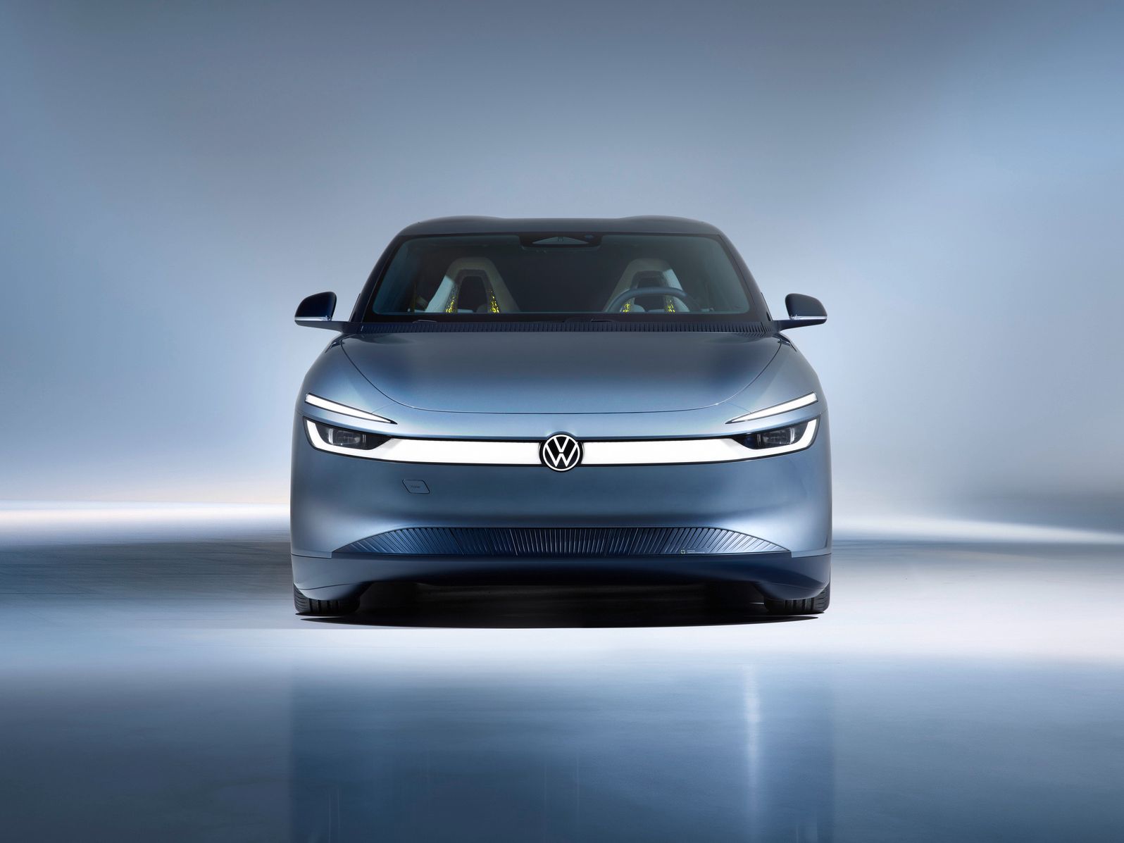 Frontansicht eines modernen, silbergrauen Elektroautos von Volkswagen in einem hell ausgeleuchteten Studio. Das Fahrzeug hat schmale, durchgehende LED-Scheinwerfer und ein minimalistisches, glattes Design. Im Innenraum sind zwei farbige Sitze schemenhaft erkennbar.