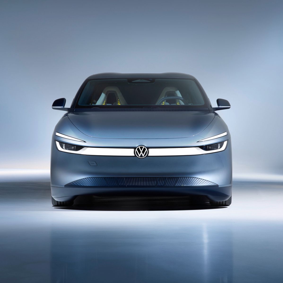 Frontansicht eines modernen, silbergrauen Elektroautos von Volkswagen in einem hell ausgeleuchteten Studio. Das Fahrzeug hat schmale, durchgehende LED-Scheinwerfer und ein minimalistisches, glattes Design. Im Innenraum sind zwei farbige Sitze schemenhaft erkennbar.