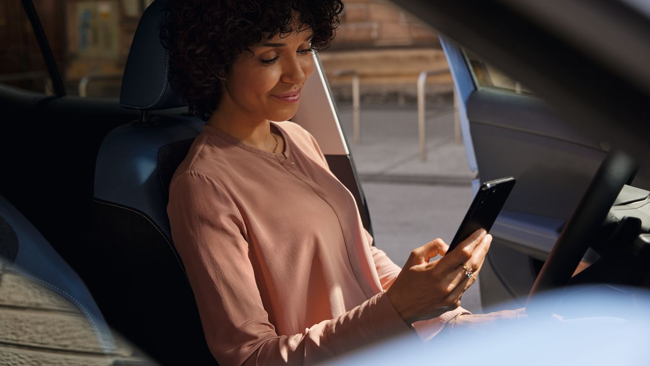 Eine lächelnde Frau schaut auf dem Fahrersitz eines geparkten VW T-Cross aufs Handy.