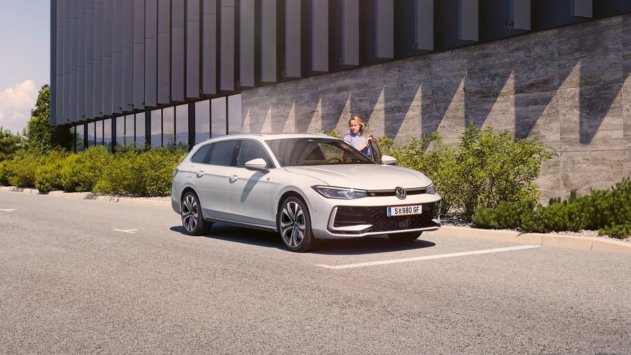  Eine Frau öffnet die Fahrertür eines weißen VW Passat R-Line und steigt ein. Im Hintergrund sieht man ein Gebäude.