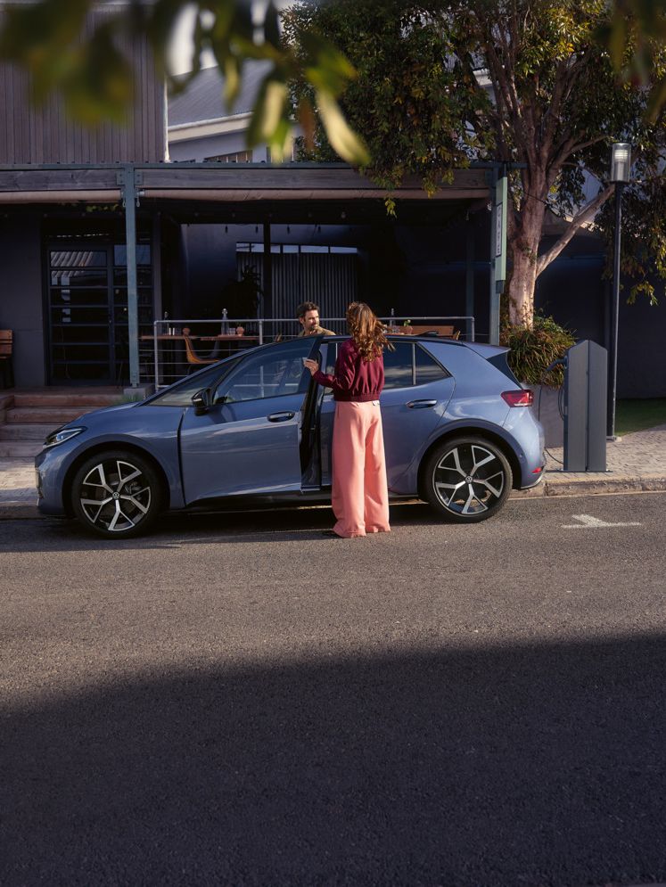 Eine Frau und ein Mann steigen in einen blauen VW ID.3 Neo, der vor einem Café parkt