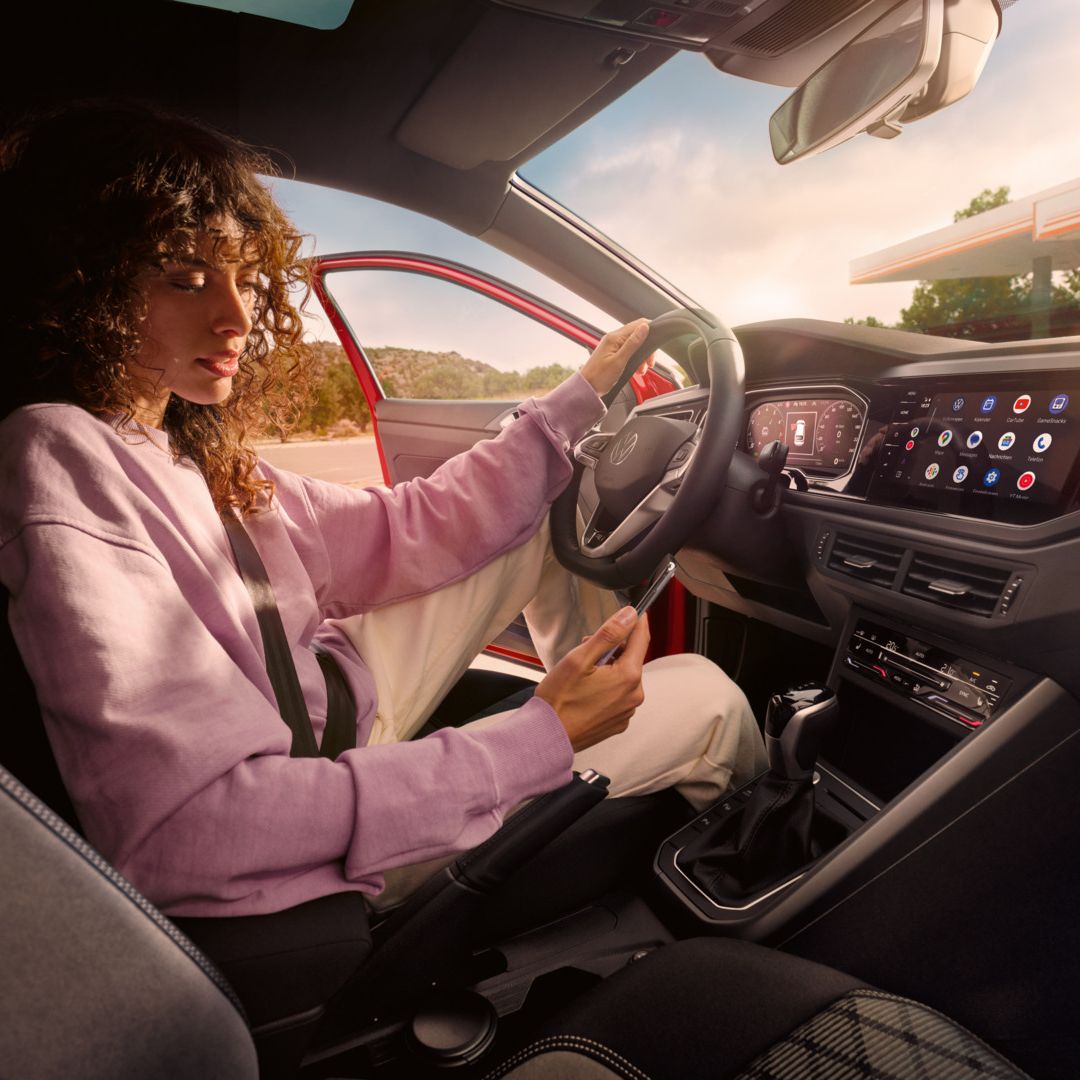 Person in einem VW Taigo sitzt auf dem Fahrersitz, trägt einen Sicherheitsgurt und hat eine Hand auf dem Lenkrad abgelegt, während sie mit der anderen ein Smartphone bedient. Auf der Mittelkonsole ist ein großes Display mit verschiedenen Apps und Funktionen zu sehen. Durch die geöffnete Fahrertür erkennt man im Hintergrund eine Tankstelle oder ein Rasthofgebäude.