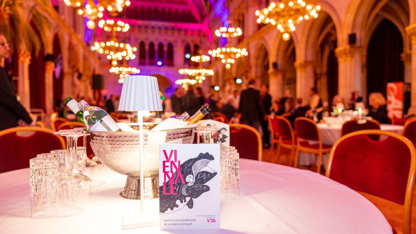 Eleganter Festsaal mit hohen Gewölbedecken und Kronleuchtern, in warmes rotes und violettes Licht getaucht. Im Vordergrund steht ein festlich gedeckter Tisch mit einem Sektkühler voller Flaschen, Gläsern und einem aufgestellten Veranstaltungsprogramm mit der Aufschrift „Wiener“. Im Hintergrund sitzen und stehen festlich gekleidete Menschen an runden Tischen.