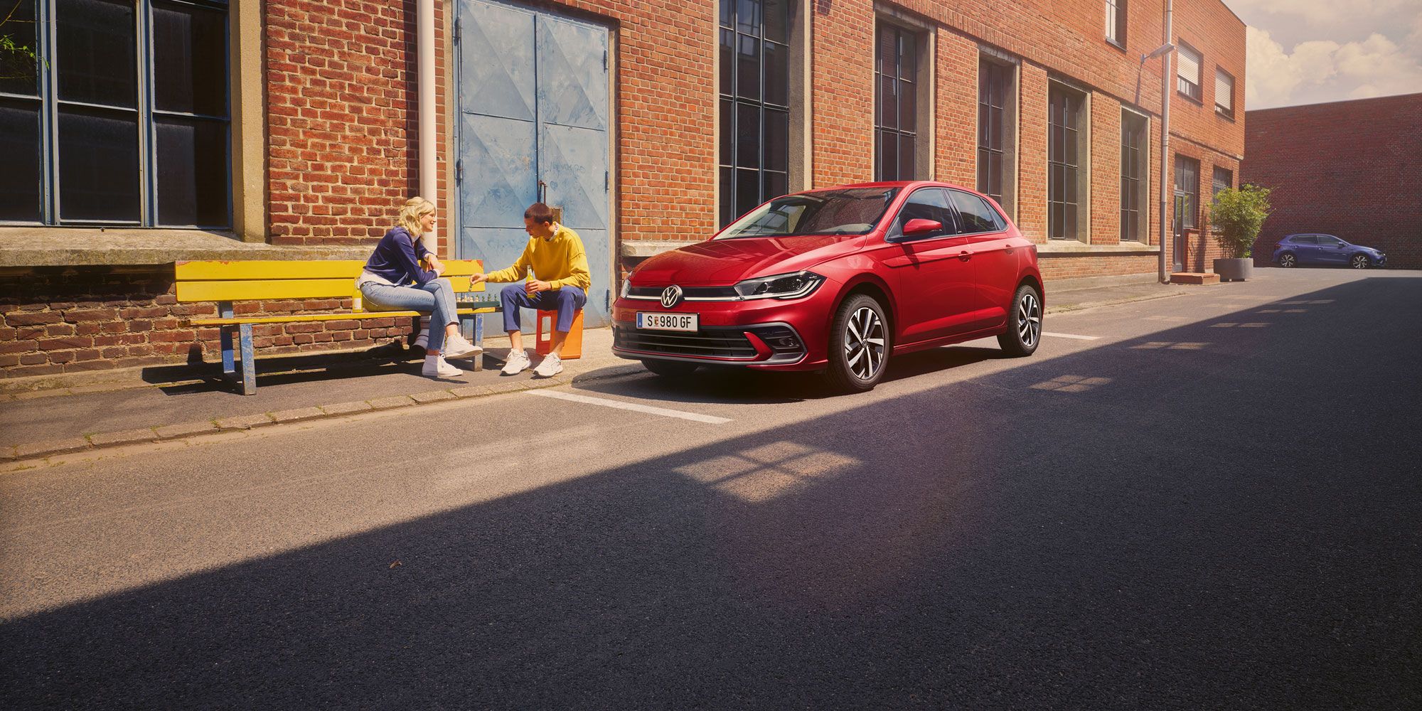 Zwei Personen spielen Schach vor einem roten VW Polo Style, der vor einem Fabrikgebäude parkt.