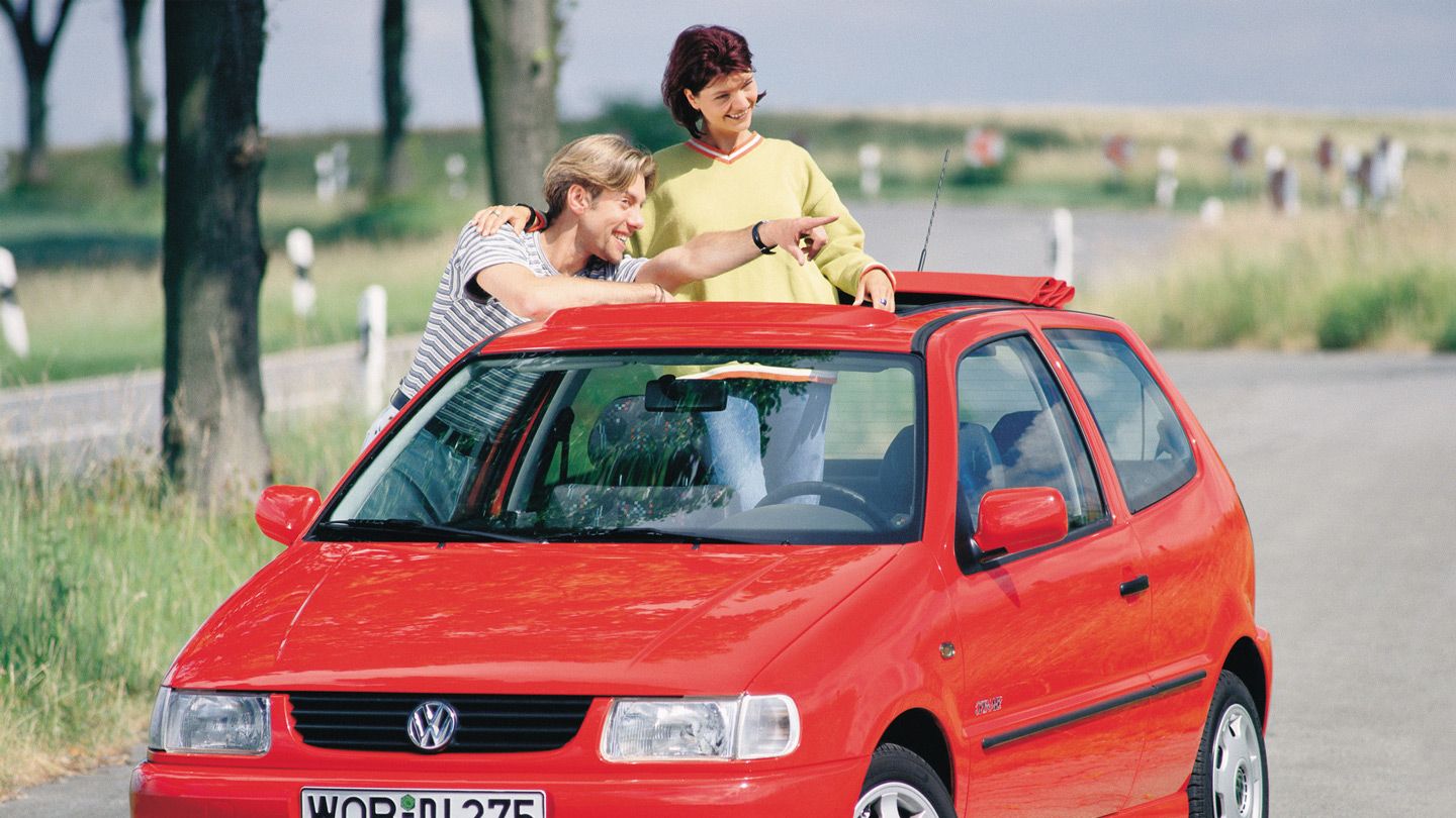 Ein roter VW Polo 3 OpenAir, geparkt auf einer Landstraße, mit zwei Personen, die mit geöffnetem Schiebedach aus dem Fahrzeug hinausschauen. Die Umgebung zeigt grüne Felder und Bäume.