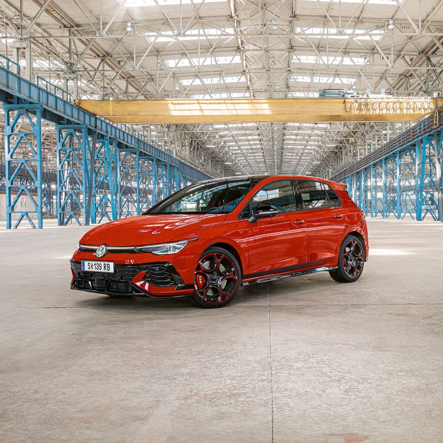 Ein roter Golf GTI steht in einer großen, leeren Industriehalle mit blauen Stahlträgern. Das Dach der Halle ist aus Glas und lässt viel Licht herein.