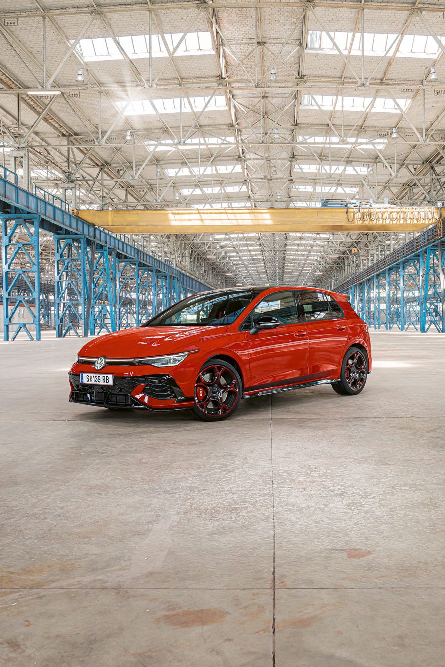 Ein roter Golf GTI steht in einer großen, leeren Industriehalle mit blauen Stahlträgern. Das Dach der Halle ist aus Glas und lässt viel Licht herein.