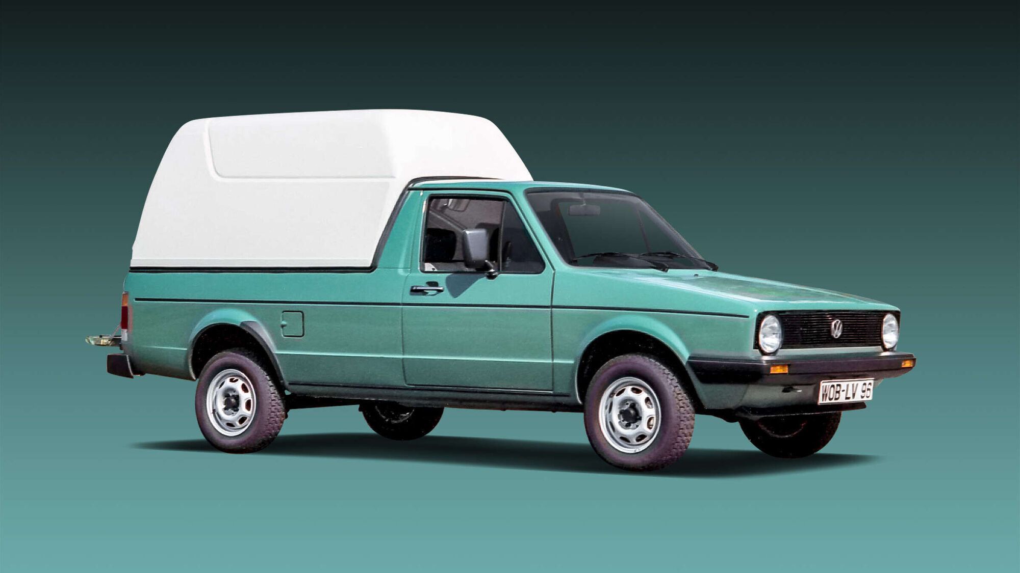 Grüner VW Caddy I mit erhöhtem weißen Aufbau auf der Ladefläche, schräg von vorne fotografiert. Das Fahrzeug steht vor einem einfarbigen, dunkel verlaufenden Hintergrund.