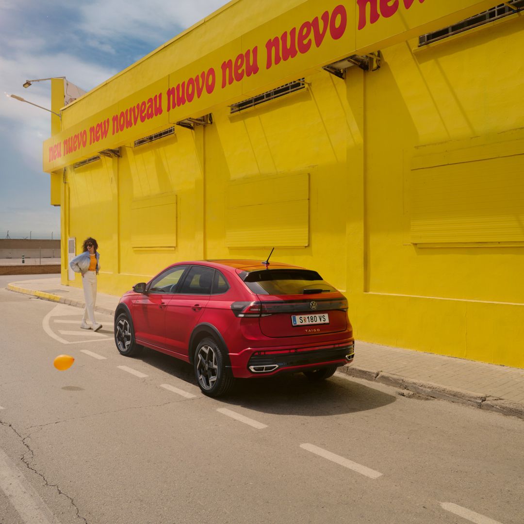 Ein roter VW Taigo steht vor einer gelben Halle am Straßenrand, eine Frau geht darauf zu.