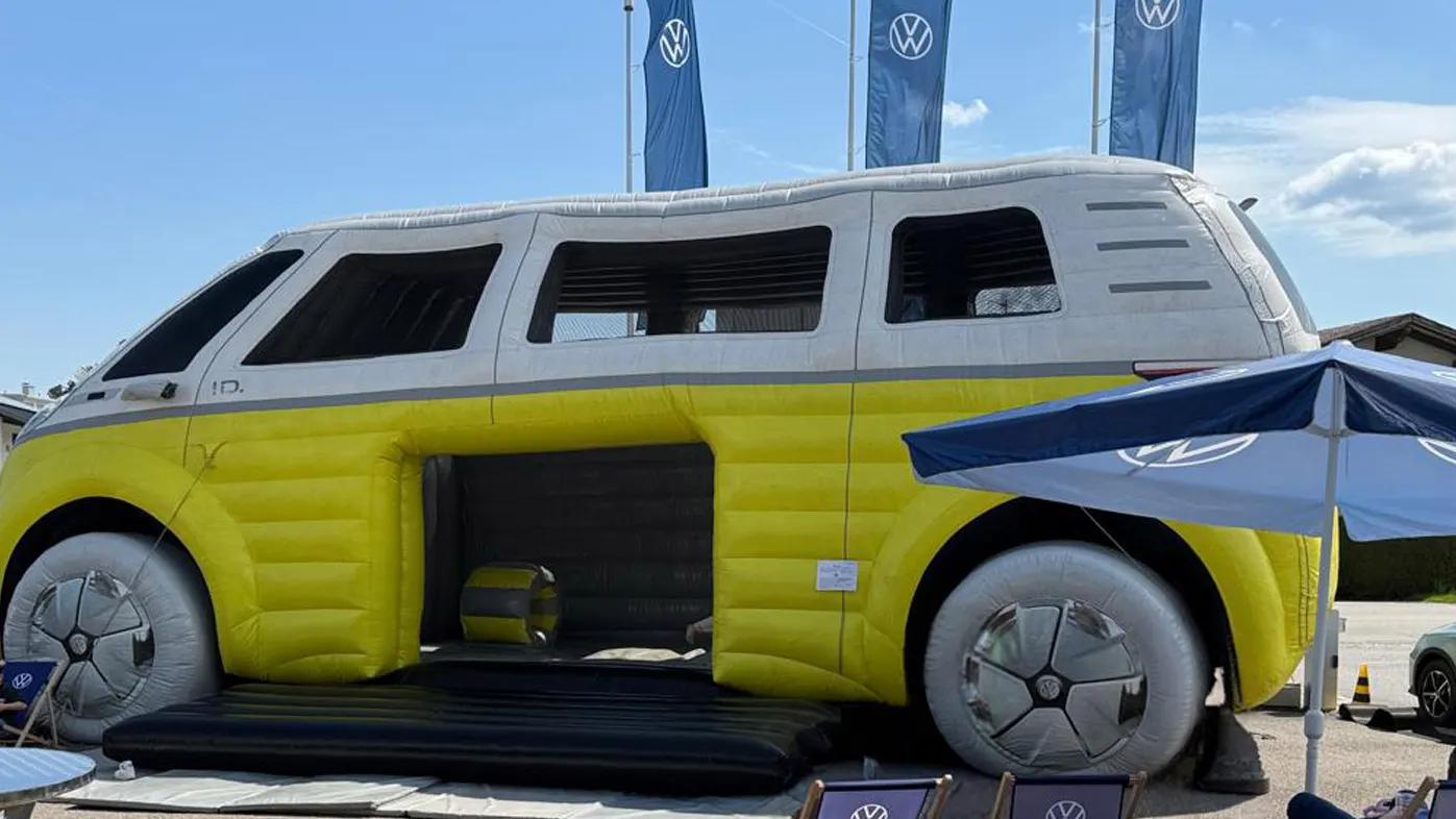 Große aufblasbare Hüpfburg in Form eines gelb-weißen VW-Bullis auf einem Freigelände bei sonnigem Wetter. Vor dem Luftfahrzeug stehen Sonnenschirme und Sitzgelegenheiten, im Hintergrund wehen mehrere blaue VW-Fahnen.