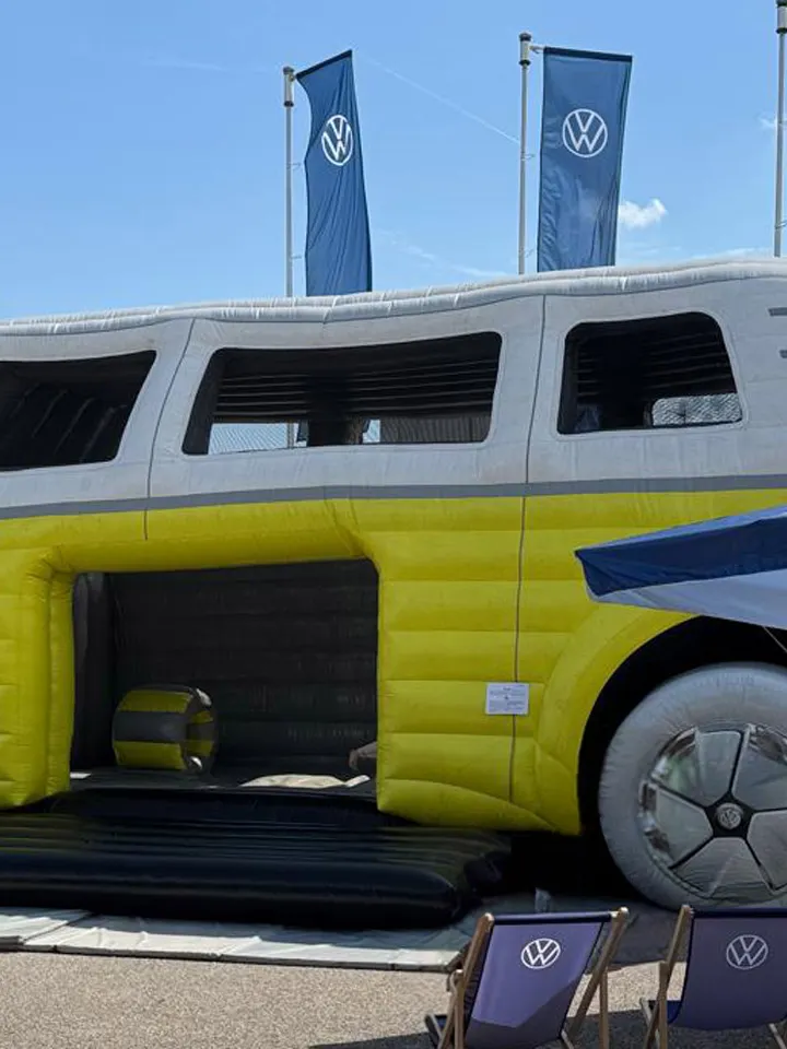 Große aufblasbare Hüpfburg in Form eines gelb-weißen VW-Bullis auf einem Freigelände bei sonnigem Wetter. Vor dem Luftfahrzeug stehen Sonnenschirme und Sitzgelegenheiten, im Hintergrund wehen mehrere blaue VW-Fahnen.