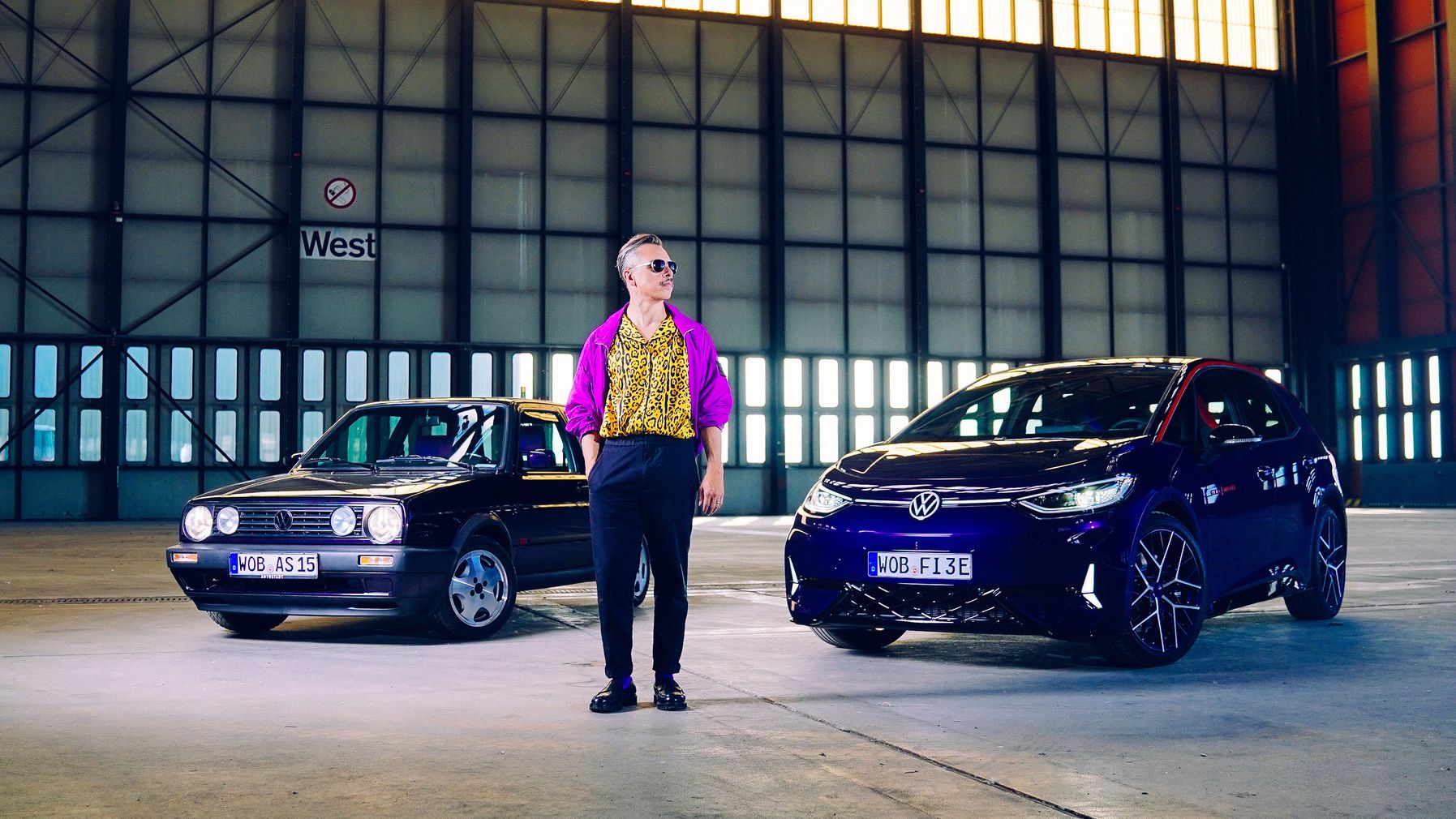 Eine Person steht in einer großen Halle zwischen einem schwarzen und einem blauen Auto. Die Autos sind von der Marke Volkswagen. Die Halle ist größtenteils leer und die Wände bestehen aus vielen Fensterpaneelen.