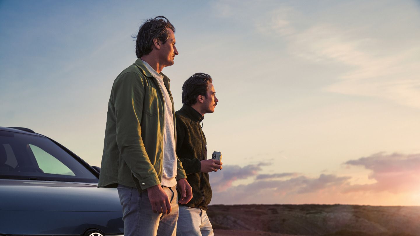 Zwei Männer lehnen an der Motorhaube eines VW ID.7 Tourer und schauen auf einen Sonnenuntergang am Meer.