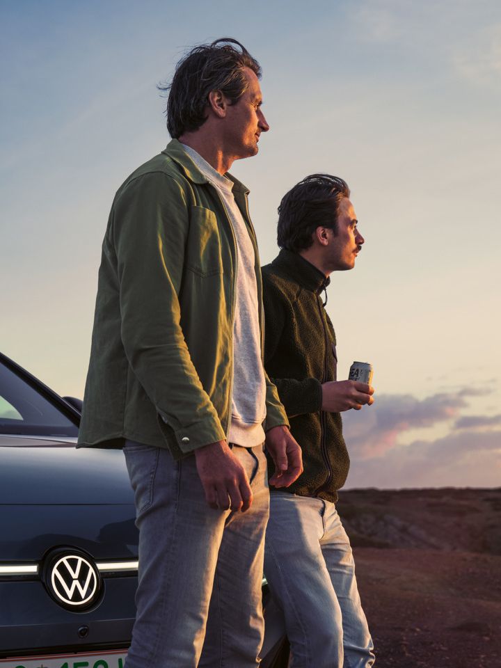 Zwei Männer lehnen an der Motorhaube eines VW ID.7 Tourer und schauen auf einen Sonnenuntergang am Meer.