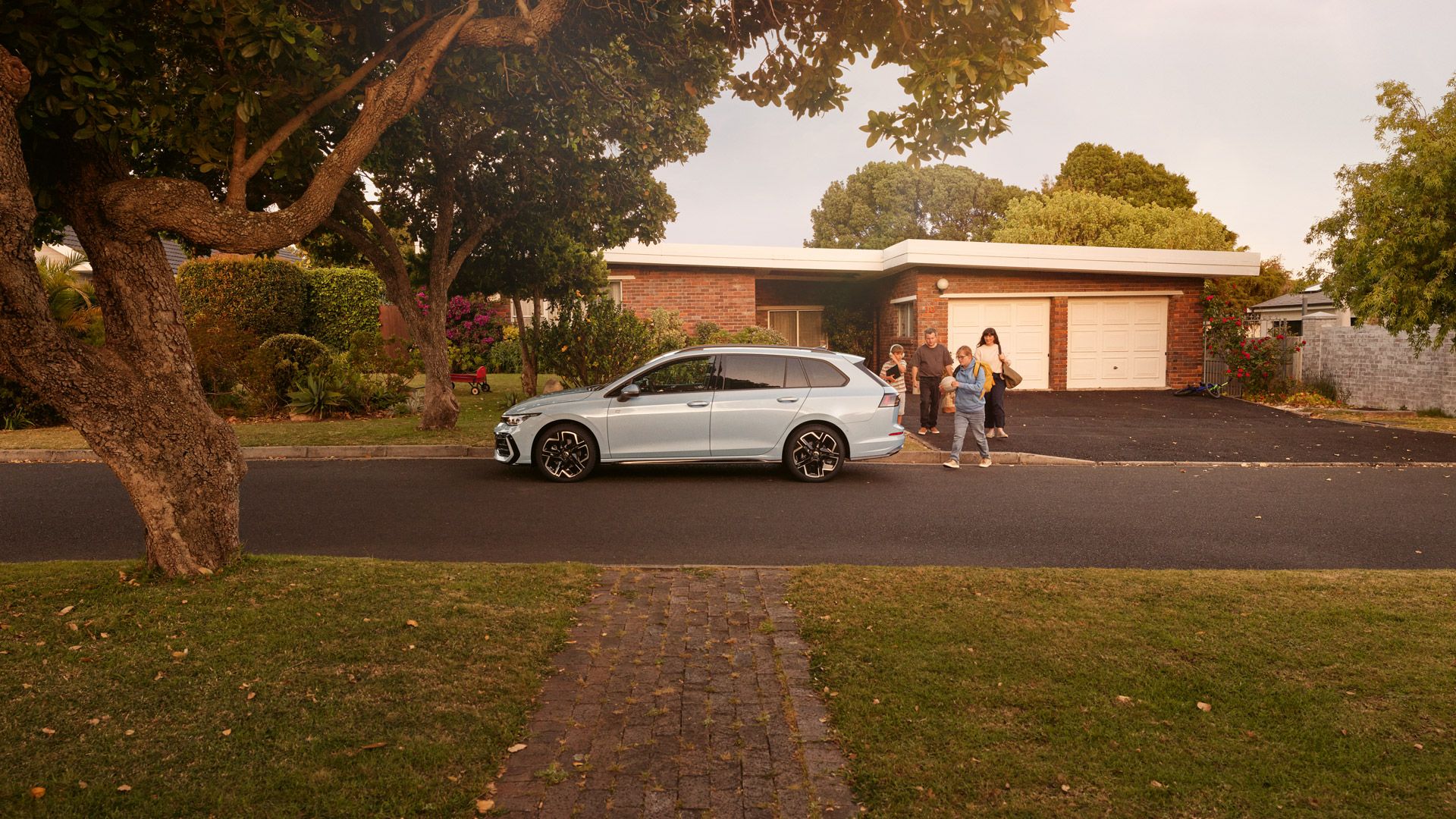 Weißer VW Golf Variant parkt in einer Wohnstraße mit Bäumen. Familie geht mit Gepäck darauf zu.