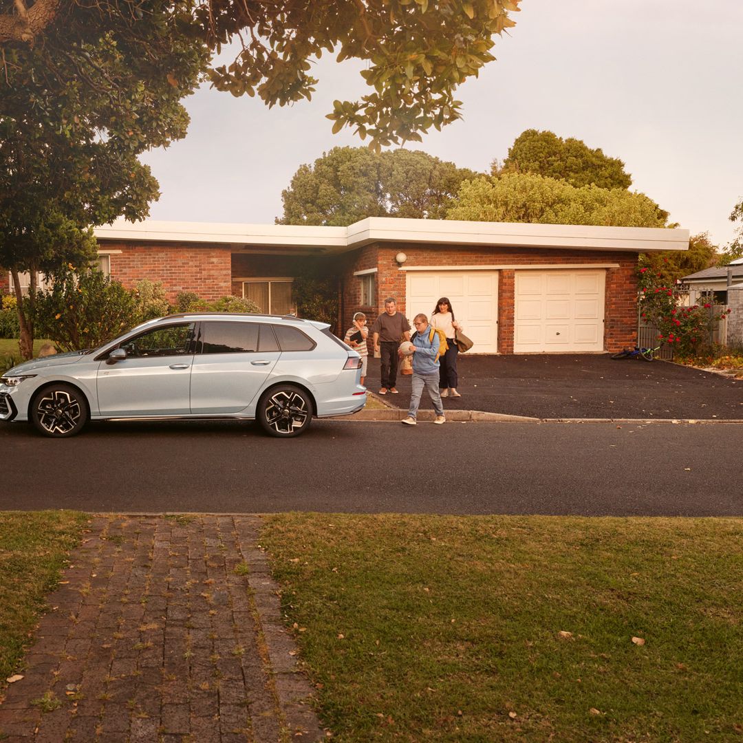 Weißer VW Golf Variant parkt in einer Wohnstraße mit Bäumen. Familie geht mit Gepäck darauf zu.