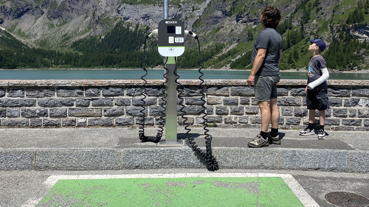 Zwei Personen stehen neben einer Ladestation für Elektrofahrzeuge in einer bergigen Landschaft mit einem See im Hintergrund. Der Ladebereich ist grün markiert und zeigt das Symbol eines Elektroautos.