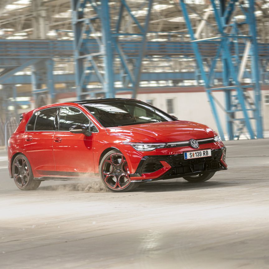 Ein roter Volkswagen Golf GTI fährt schnell durch eine leere Industriehalle. Staub wird von den Reifen aufgewirbelt. Im Hintergrund sind blaue Stahlkonstruktionen und Fenster zu sehen.