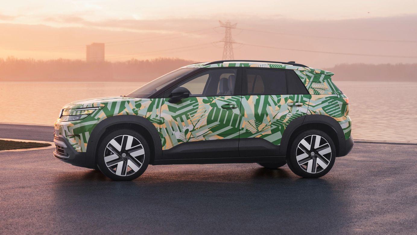 Ein SUV mit einer grünen und weißen Tarnlackierung steht auf einem asphaltierten Platz vor einem Gewässer bei Sonnenuntergang. Im Hintergrund sind Strommasten und Gebäude zu sehen. Der Himmel ist leicht bewölkt und in warmen Farben gehalten.