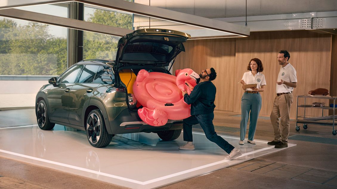 Ein Mann drückt einen aufblasbaren Flamingo in den offenen Kofferraum des VW ID. Cross. Zwei Personen stehen daneben und gucken ihm amüsiert zu.