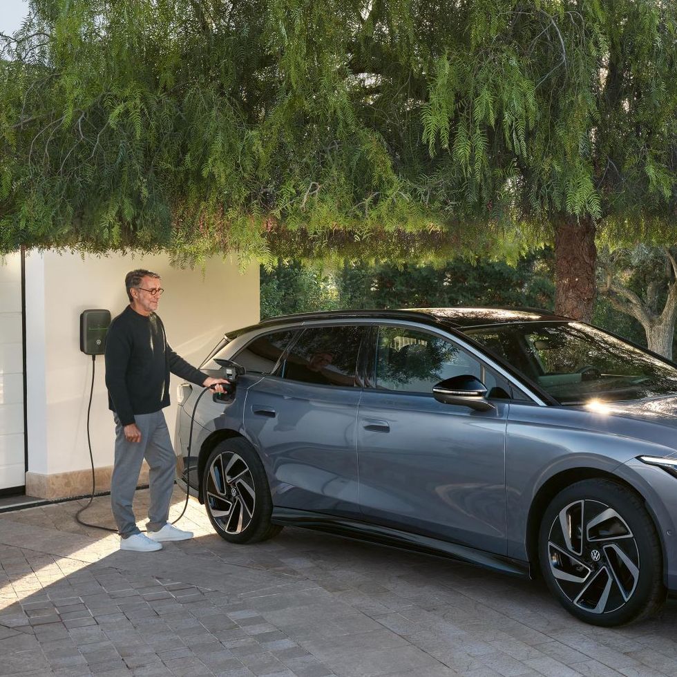 Ein Mann steht neben einem blauen VW ID.7 Tourer, der vor einer Garage an einer Wallbox lädt.