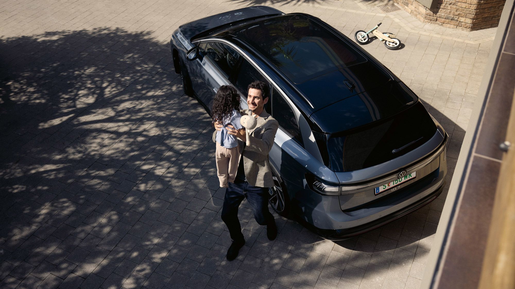 Mann hebt sein Kind hoch, was eine Puppe in der Hand halt und dahinter steht geparkt in der Einfahrt ein ID.7 Tourer und ein umgekipptes Kinderfahrrad.