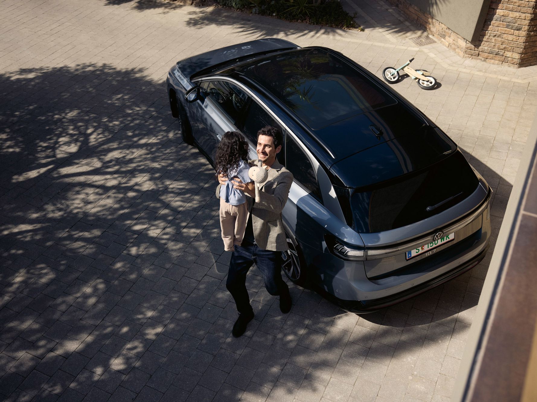 Mann hebt sein Kind hoch, was eine Puppe in der Hand halt und dahinter steht geparkt in der Einfahrt ein ID.7 Tourer und ein umgekipptes Kinderfahrrad.