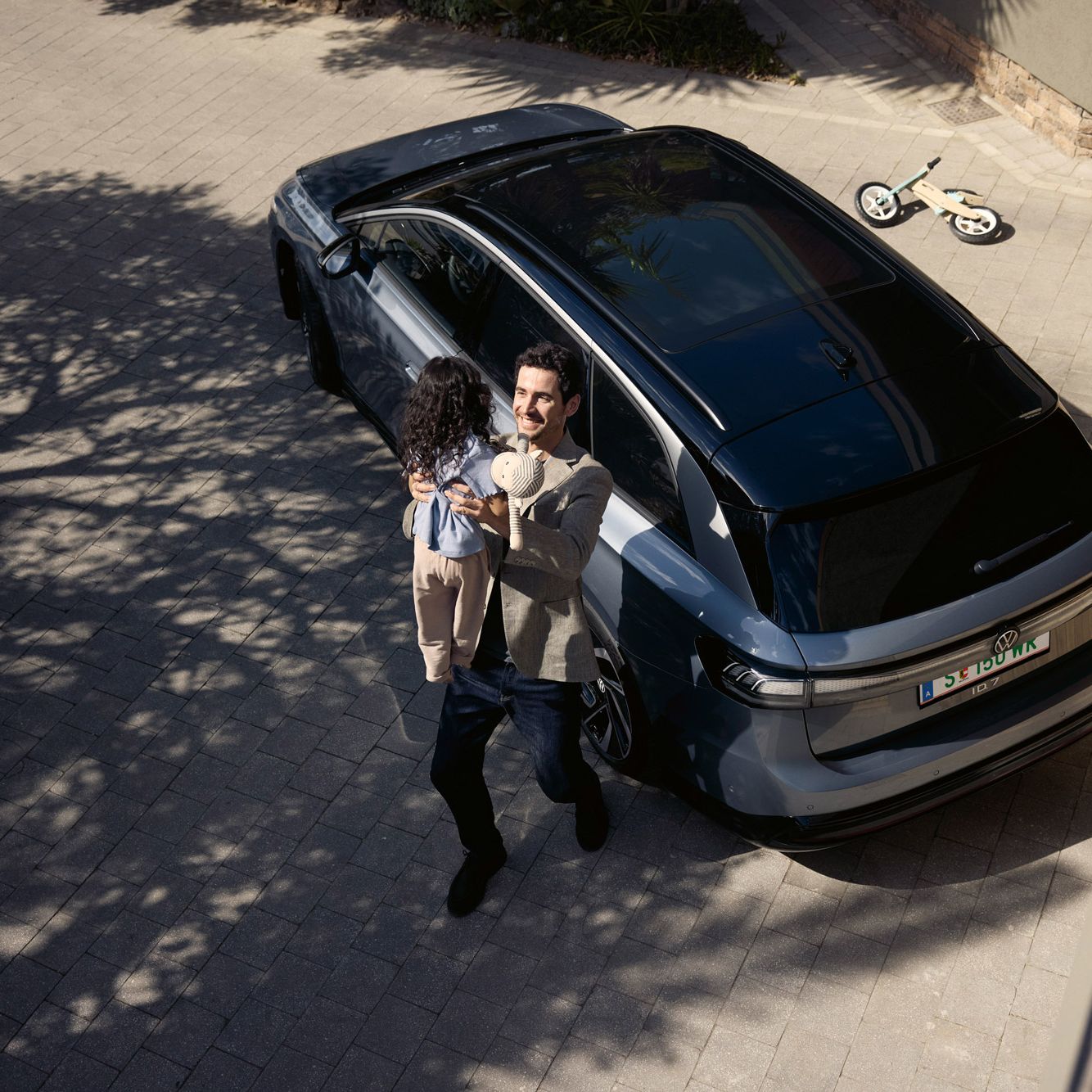 Mann hebt sein Kind hoch, was eine Puppe in der Hand halt und dahinter steht geparkt in der Einfahrt ein ID.7 Tourer und ein umgekipptes Kinderfahrrad.