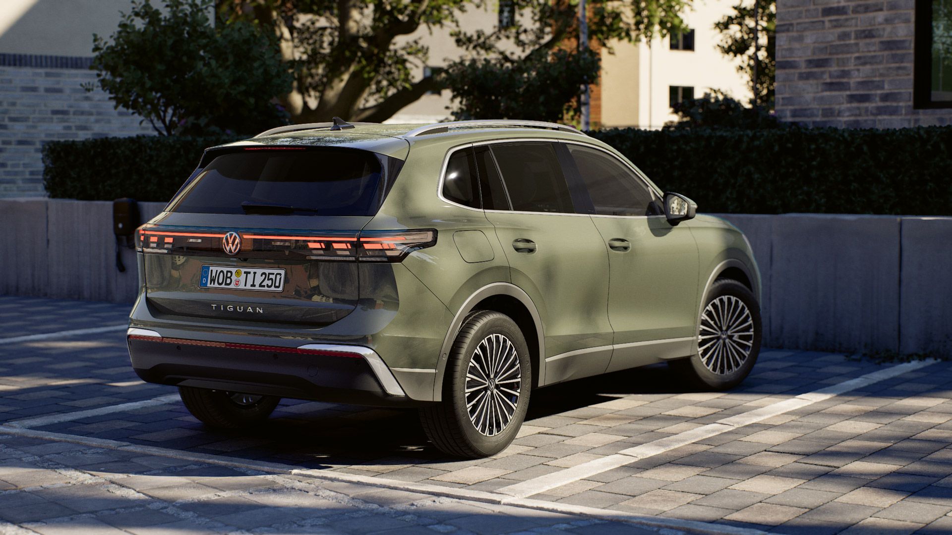 Ein olivgrüner Volkswagen Tiguan steht auf einem gepflasterten Parkplatz neben einem modernen Gebäude. Das Fahrzeug ist von hinten schräg zu sehen, mit LED-Rückleuchten und getönten Scheiben. Im Hintergrund sind eine niedrige Mauer, Sträucher und Bäume in der Sonne zu erkennen.