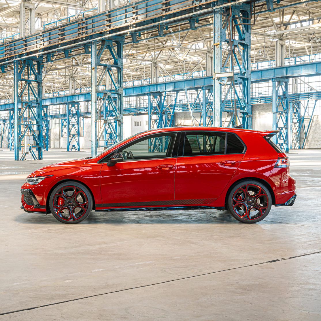 Ein roter Golf GTI steht in einer großen, leeren Industriehalle mit blauen Stahlträgern. Die Halle hat ein hohes Dach und ist gut beleuchtet. Es gibt viel Platz um das Fahrzeug herum.