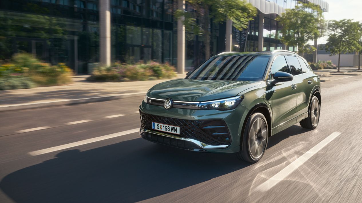 Ein Tiguan eHybrid in Cipresso-Grün fährt auf der Straße. Im Hintergrund sind ein modernes Gebäude und Bäume zu sehen. 