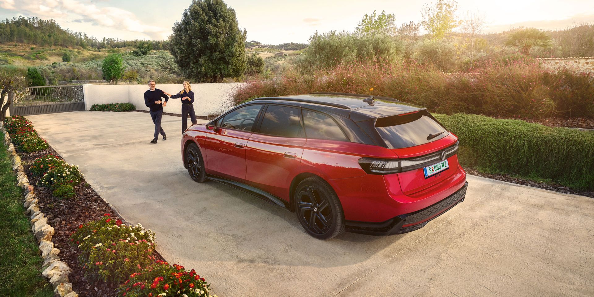 Ein roter VW ID.7 GTX Tourer parkt in einer langen Auffahrt, ein Mann und eine Frau wollen einsteigen.