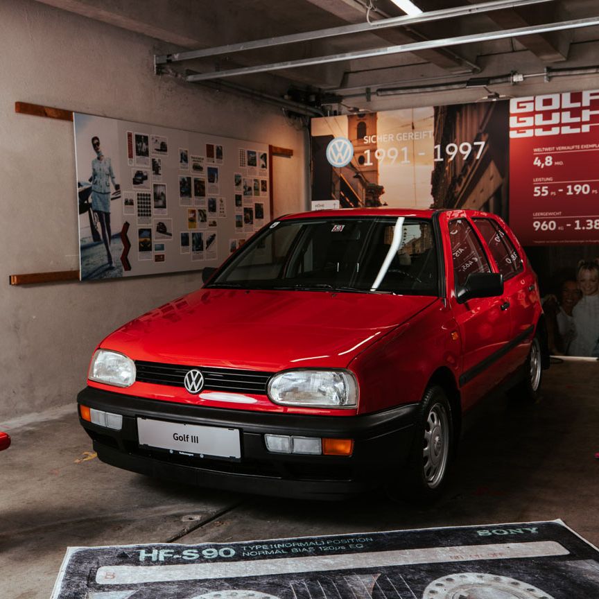 Ein roter VW Golf 3 steht in einer Garage, umgeben von Postern und Autoteilen. Auf einem Poster wird ein historischer Überblick über verschiedenen Golf-Modelle gezeigt. Der Boden der Garage ist mit Werkzeugen und Teile bedeckt.