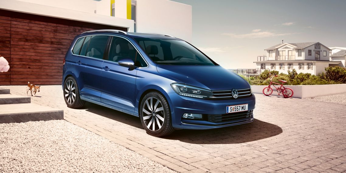 Blauer VW Touran steht vor einem modernen Einfamilienhaus, eine Familie geht darauf zu.