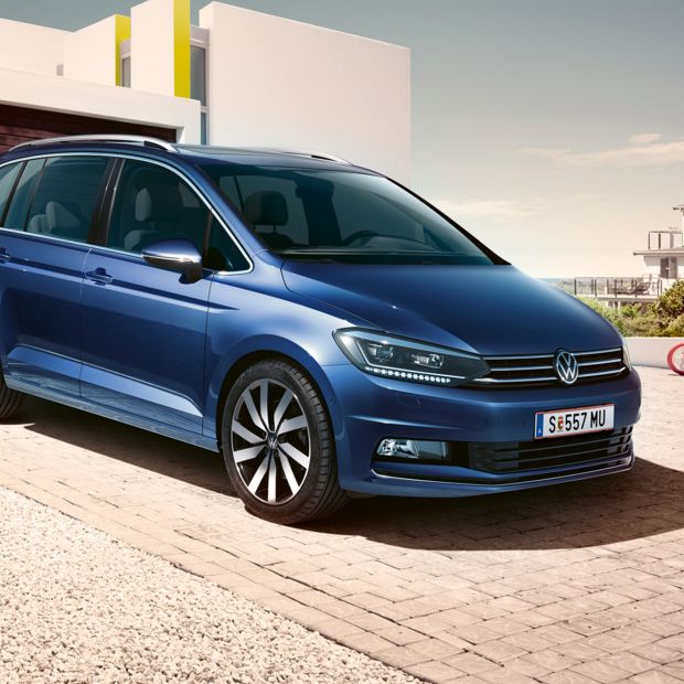 Blauer VW Touran steht vor einem modernen Einfamilienhaus, eine Familie geht darauf zu.