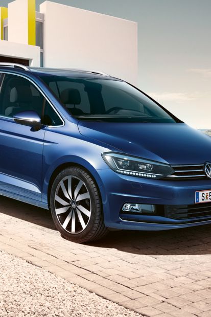 Blauer VW Touran steht vor einem modernen Einfamilienhaus, eine Familie geht darauf zu.