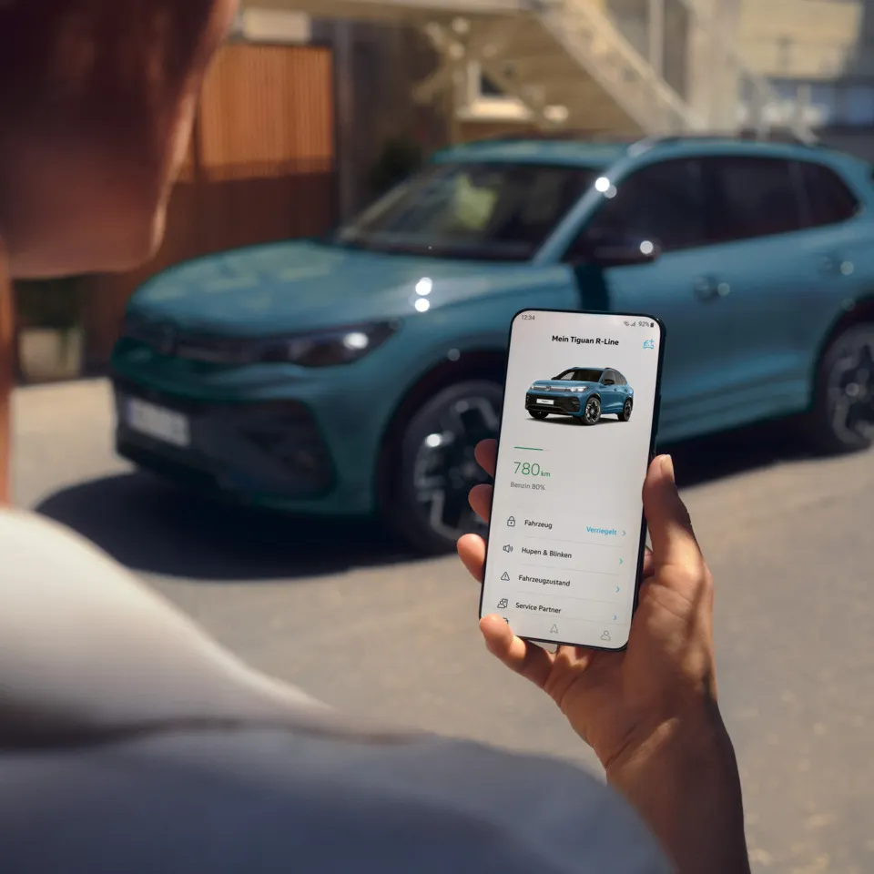  Blick auf ein Handydisplay mit Fahrzeugdaten des Tiguan, im Hintergrund ein seitlich geparkter VW Tiguan in blau.
