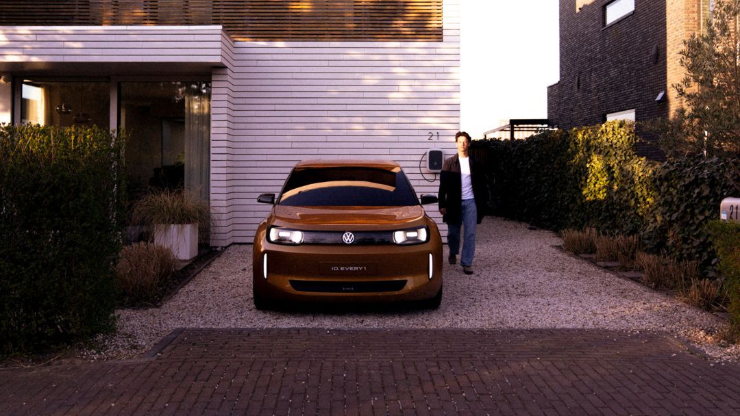 Moderner, zweistöckiger Kubusbau mit heller Fassade und großen Holzlamellen, vor dem ein goldbraunes Elektroauto in einer Kiesauffahrt parkt. Rechts neben dem Auto steht eine Person in Freizeitkleidung und blickt zum Haus. Die Umgebung wirkt wie ein gehobenes Wohnviertel mit gepflegten Hecken und Nachbarhäusern.