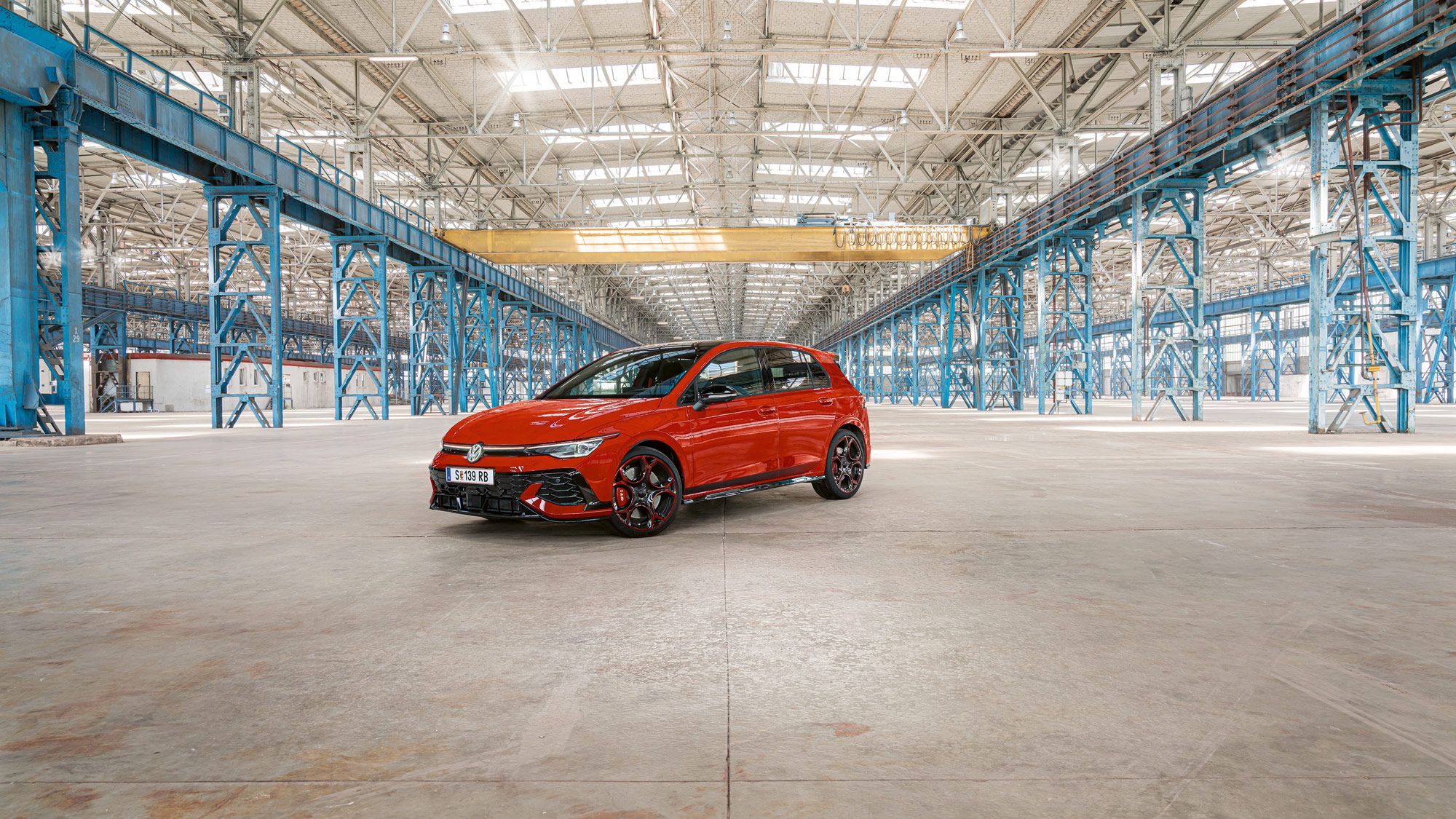 Ein roter Golf GTI steht in einer großen, leeren Industriehalle mit blauen Stahlträgern. Das Dach der Halle ist aus Glas und lässt viel Licht herein.