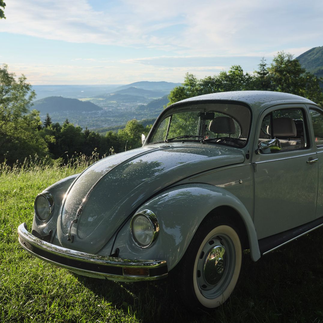 Ein grauer VW Käfer steht auf einer grünen Wiese. Im Hintergrund sind Bäume und hügelige Landschaften unter einem bewölkten Himmel zu sehen. Die Sonne scheint durch die Wolken und beleuchtet das Auto.