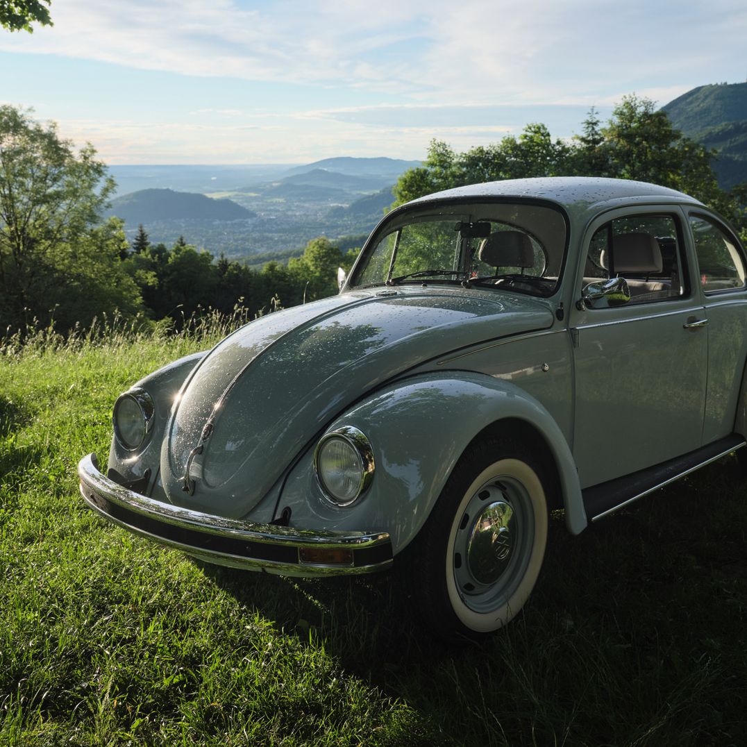 Ein grauer VW Käfer steht auf einer grünen Wiese. Im Hintergrund sind Bäume und hügelige Landschaften unter einem bewölkten Himmel zu sehen. Die Sonne scheint durch die Wolken und beleuchtet das Auto.