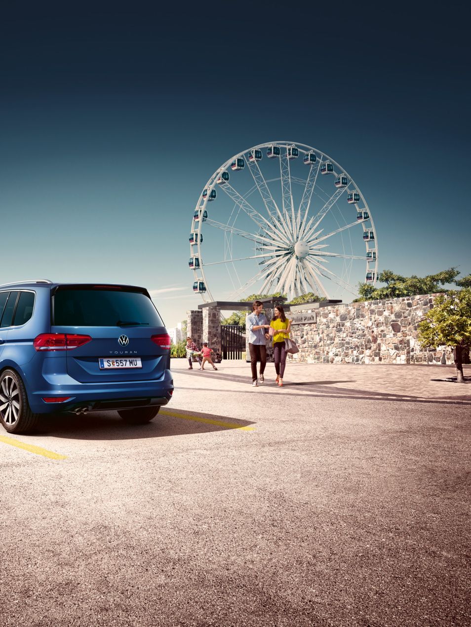 Touran steht am Parkplatz, Familie mit zwei Kindern auf dem Weg zum Auto, im Hintergrund ein Riesenrad.