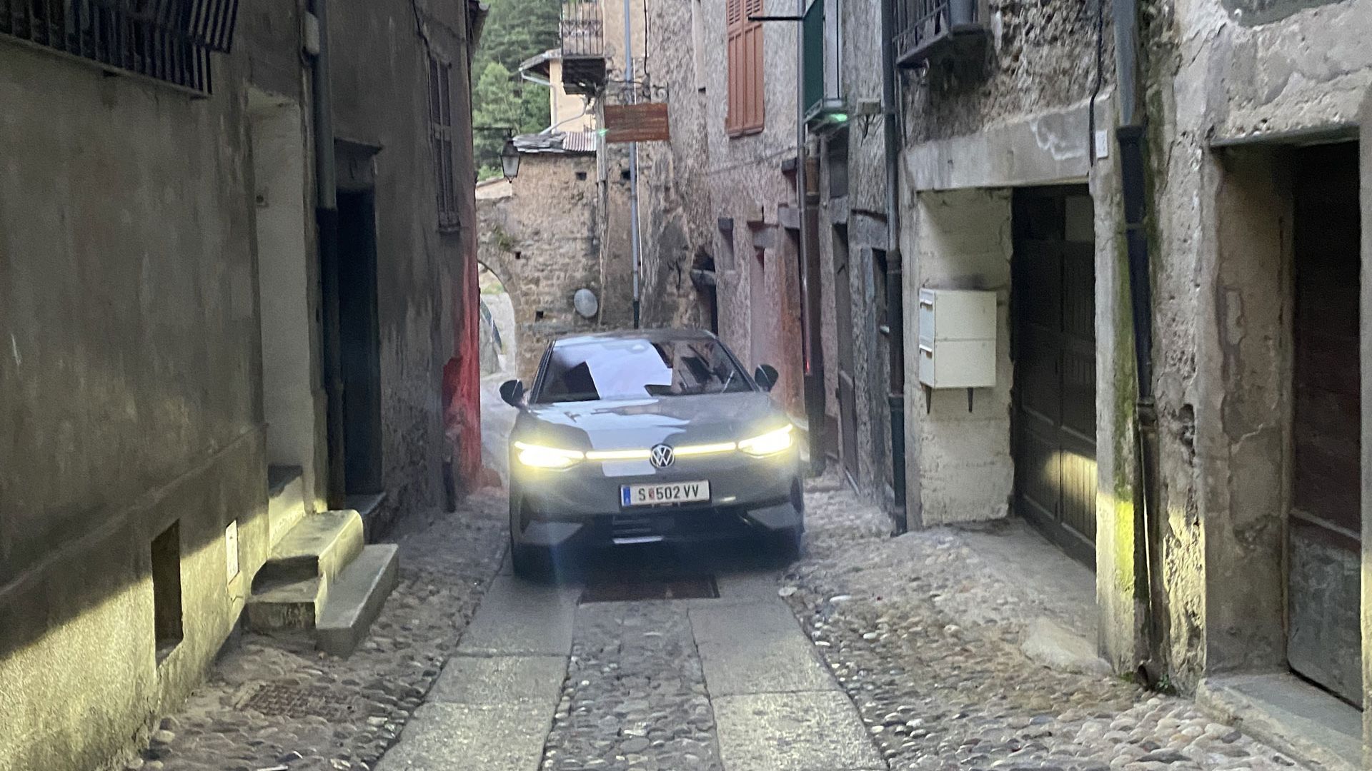Ein modernes Auto fährt durch eine enge, steinige Gasse in einer historischen Stadt. Die Straße ist von alten Gebäuden mit Steinwänden und Holztüren gesäumt. Der Weg ist so schmal, dass das Auto nur knapp hindurch passt.